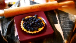 Une tartelette à la pâte dorée, garnie d'une crème jaune et entièrement recouverte de myrtilles sombres, posée sur une petite assiette carrée bordeaux. Un rouleau à pâtisserie en bois traverse l'arrière-plan, sur un linge foncé.
