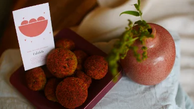 Des falafels ronds et dorés, à la surface rugueuse, disposés sur une assiette carrée bordeaux. À côté, une carte crème illustrée d'un bol corail annonce « Falafels », près d'un petit vase rond rose d'où s'échappe une tige verte.