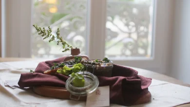 Une planche en bois dressée devant une fenêtre : un bocal en verre garni de tartare d'algues et de pousses vertes, un linge bordeaux froissé et une brindille fleurie. La lumière du jour traverse la scène à contre-jour.