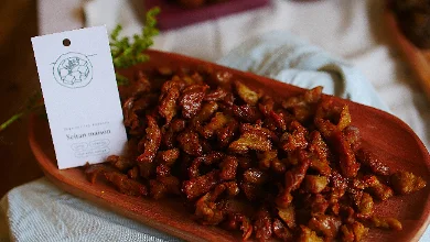 Des lamelles de seitan caramélisées, brunes et brillantes, servies généreusement dans un plat ovale en bois sombre. Une étiquette crème au tampon rond indique « Seitan maison », posée contre le plat sur un linge clair.