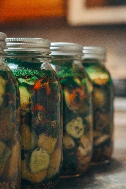Une rangée de grands bocaux en verre remplis de rondelles de concombre, de lamelles de poivron rouge et de brins d'aneth, alignés dans une pièce à la lumière chaude et tamisée.
