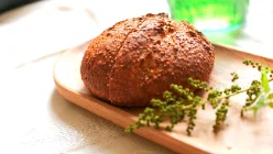 Une miche de pain ronde à la croûte brune et farinée, grignée d'une croix sur le dessus, posée sur une planche en bois clair avec un brin d'herbe fleurie. En arrière-plan flou, un verre vert capte la lumière.