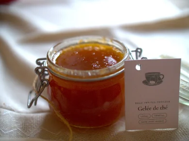 Un petit bocal à joint rempli d'une gelée ambrée et translucide, posé sur un tissu blanc froissé. Une étiquette rose pâle, illustrée d'une tasse à thé, indique « Gelée de thé » sous le nom Deux Petites Pousses.