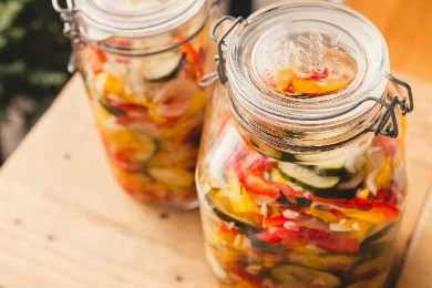Deux grands bocaux en verre à joint, remplis de légumes marinés en lamelles — courgette, poivron rouge et jaune, oignon — posés sur une planche en bois clair dans une lumière douce.